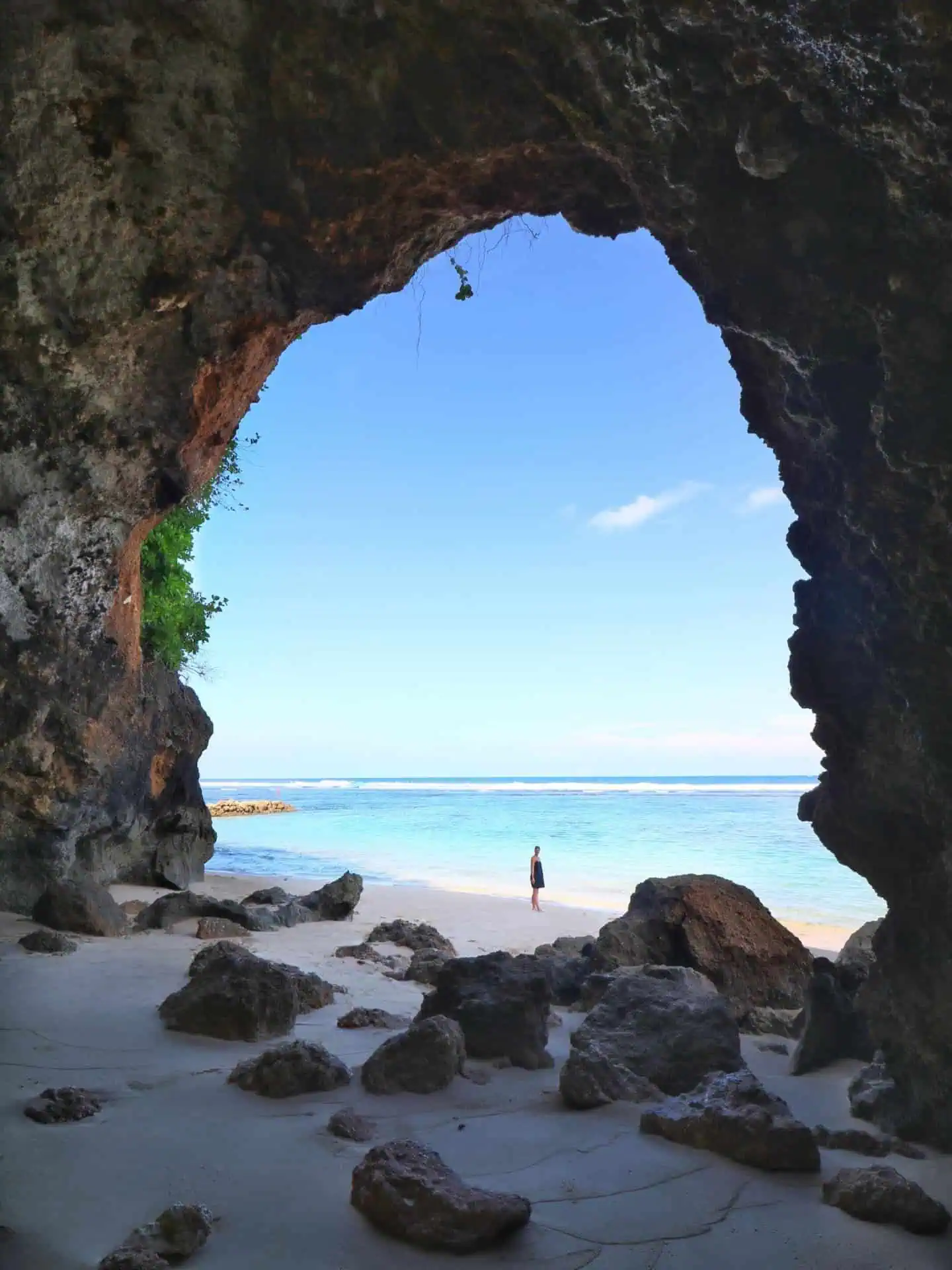 Beautiful round-shaped gunung payung cave with a woman standing inside, clear blue water and blue sky in the background