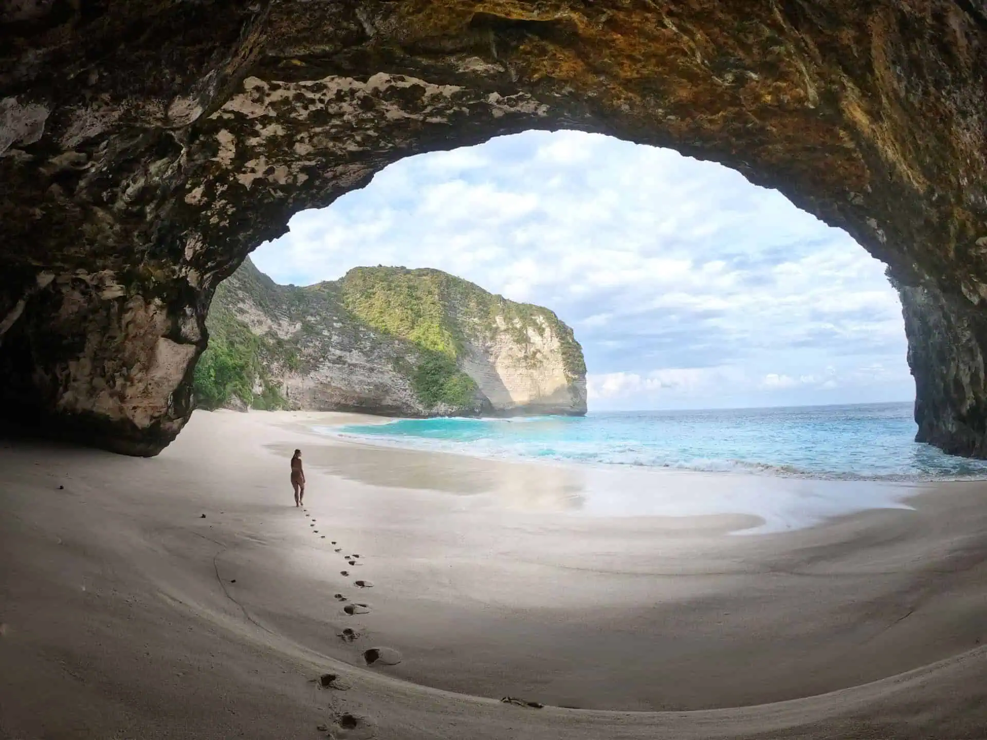 Woman walking out of Kelingking Beach Cave with light clouds, blue sea and a big rock in the background