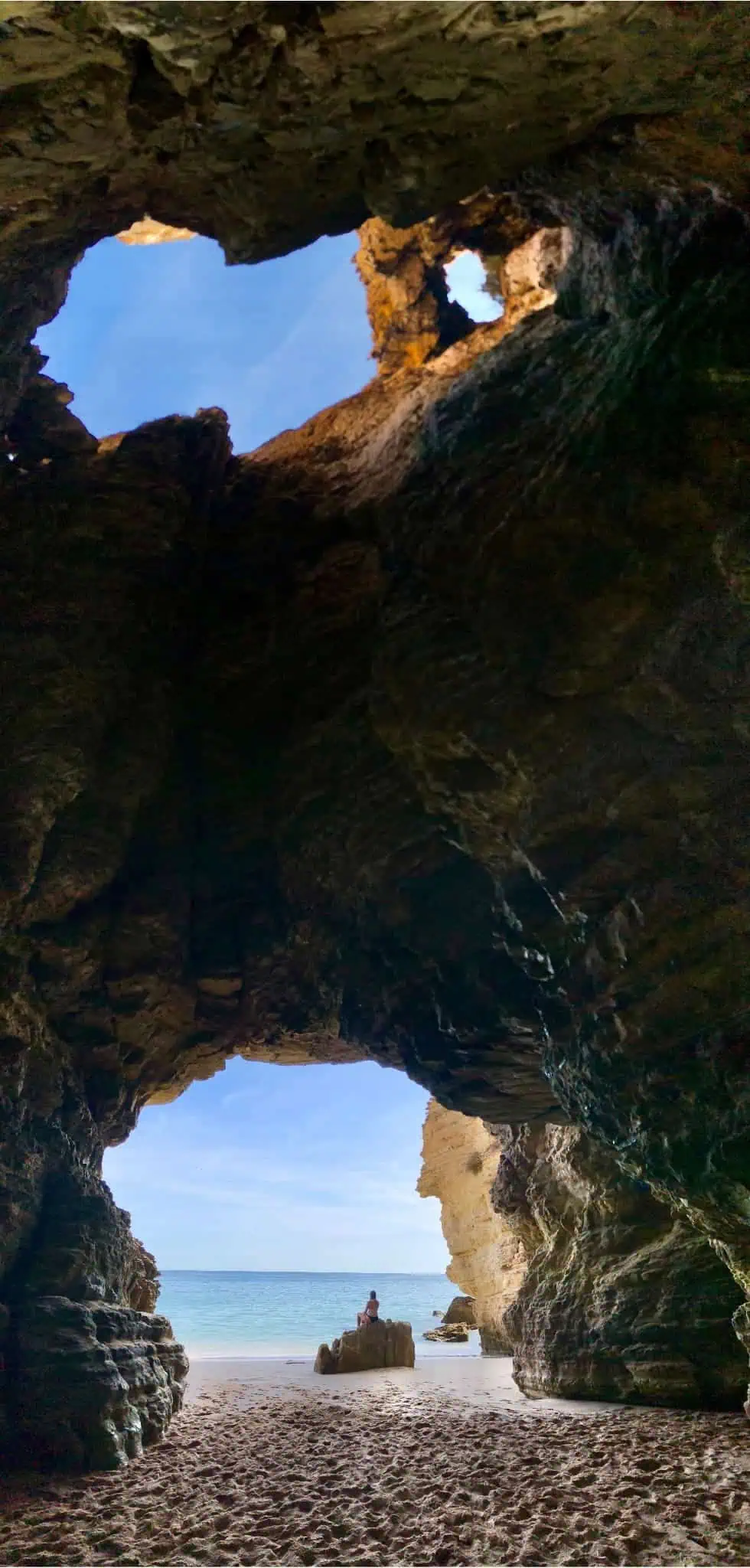 Vertical panoramic view of the cave at the very end of praia do beliche showing the to ceiling hole and the entrance where a woman is sitting on a rock