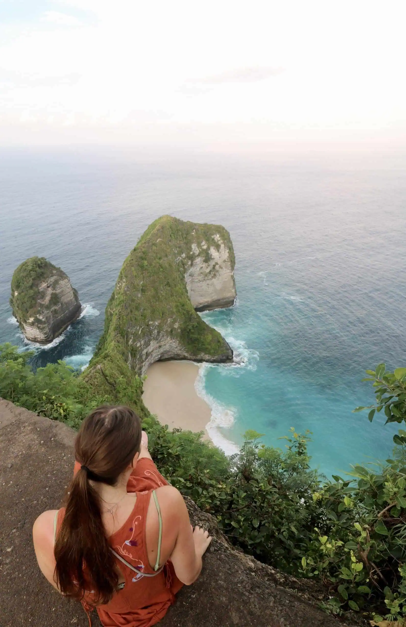 A woman sitting in front of the dinosaur shaped cliff of Kelingking beach with the surrounding clear blue waters