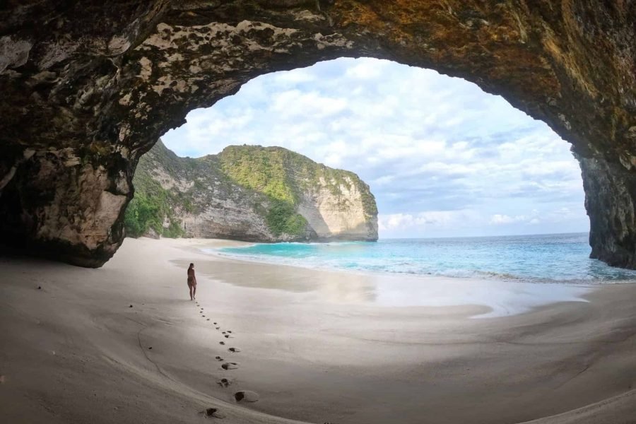 Woman walking out of Kelingking Beach Cave with light clouds, blue sea and a big rock in the background