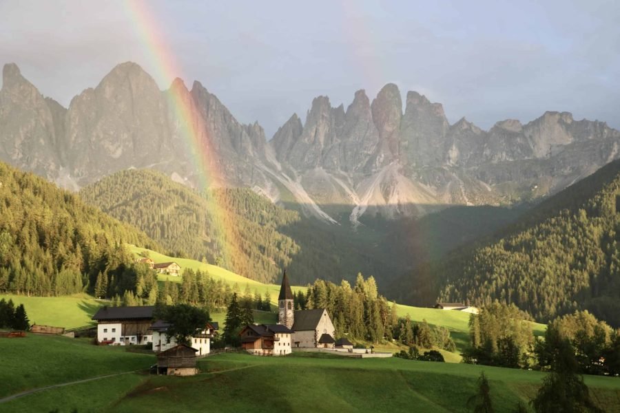 Rainbow over Santa Maddalena's quaint village, with the famous church surrounded by dark forest and the Dolomite mountains in the background, Val di Funes, Italy