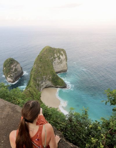 A woman sitting in front of the dinosaur shaped cliff of Kelingking beach with the surrounding clear blue waters